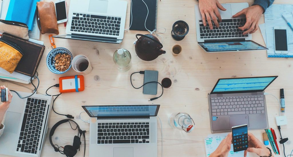 two people working on four laptops that are sitting on a large wooden desk and are surrounded by notebooks