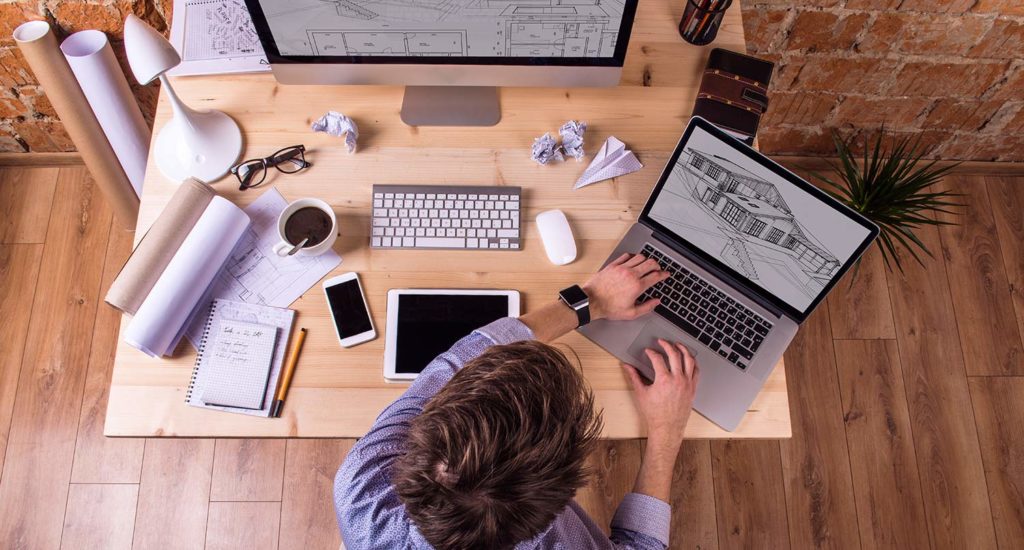 Man working on a laptop on a wooden desk with a tablet and desktop computer