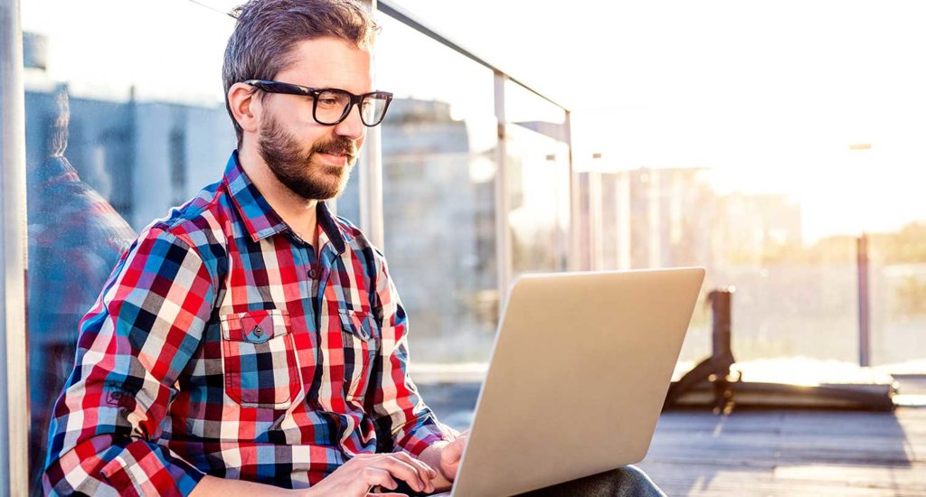 Man sitting on the roof of his apartment building tapping on a silver laptop