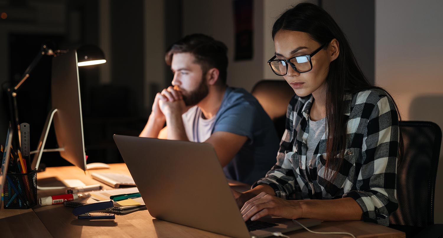 woman working on a laptop creating an ecommerce website at night with a man working on a desktop computer behind her