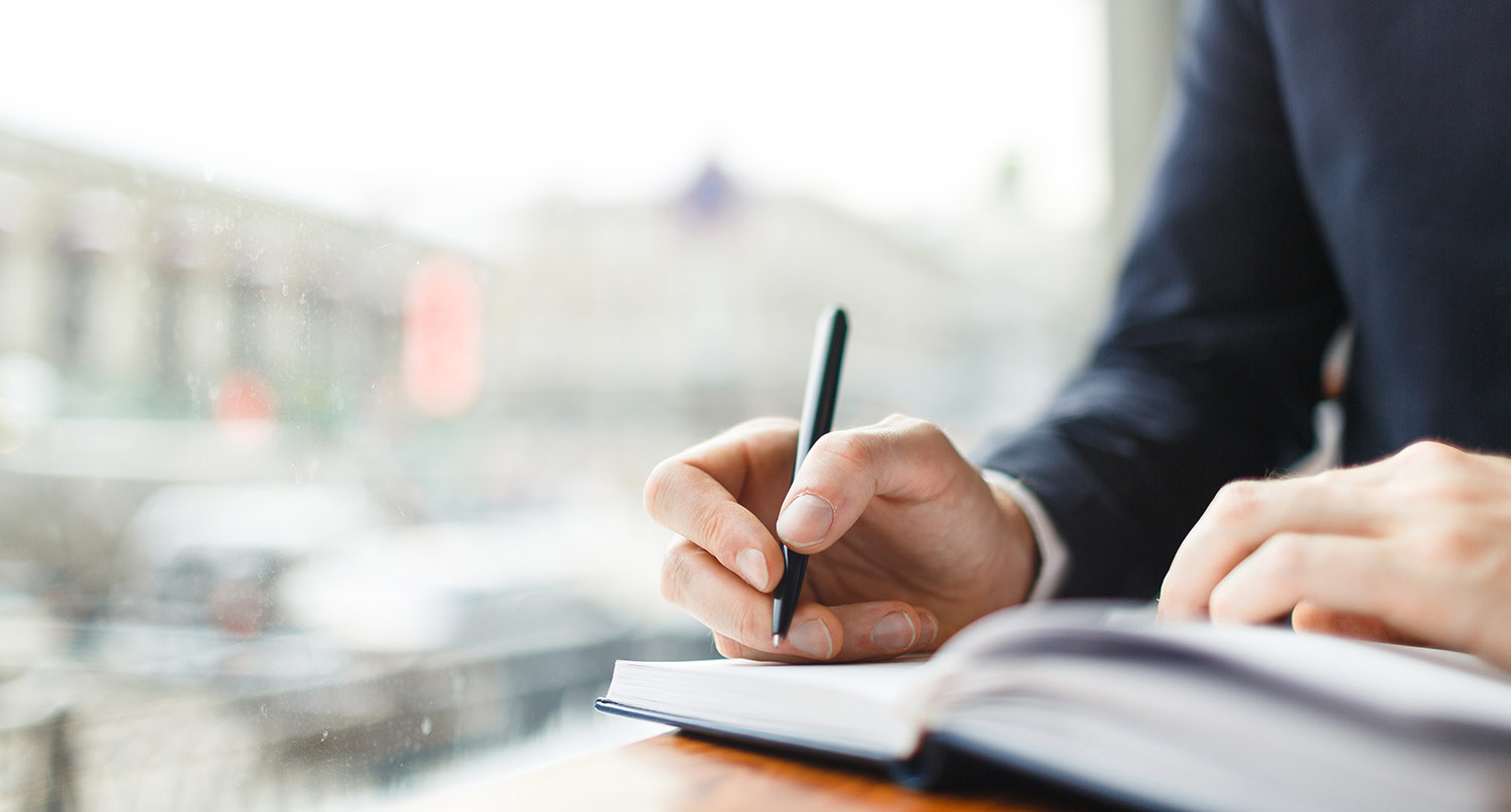 man in business suit writing into a notebook next to a window at a coffee shop