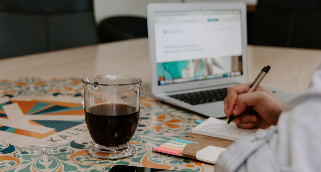 a person writing in a journal with a glass of a beverage on their left and a laptop with their personal website on the right