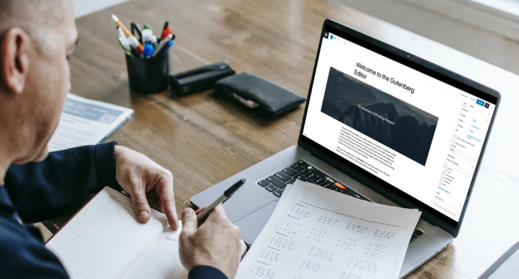 A man writing in a notebook with a laptop with the WordPress Gutenberg editor showing