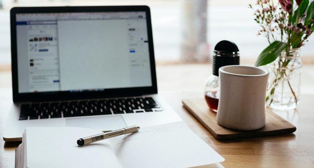 A notebook and pen sitting on a desk next to a laptop and a cup of tea