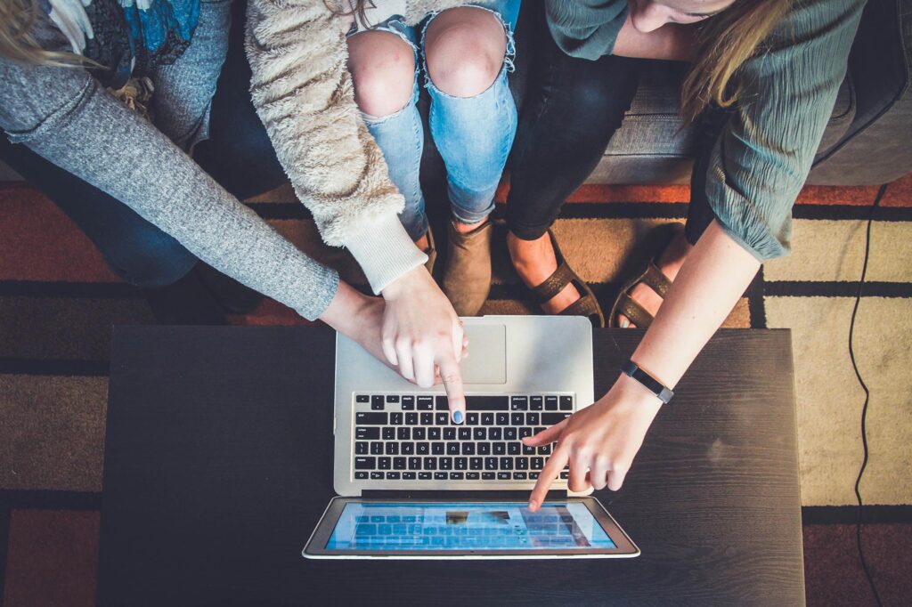 three people sitting on a couch looking and pointing at a laptop sitting on a wooden table