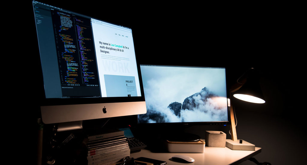 an iMac showing code and a website and another monitor with a basic background sitting on a white desk in a dark room