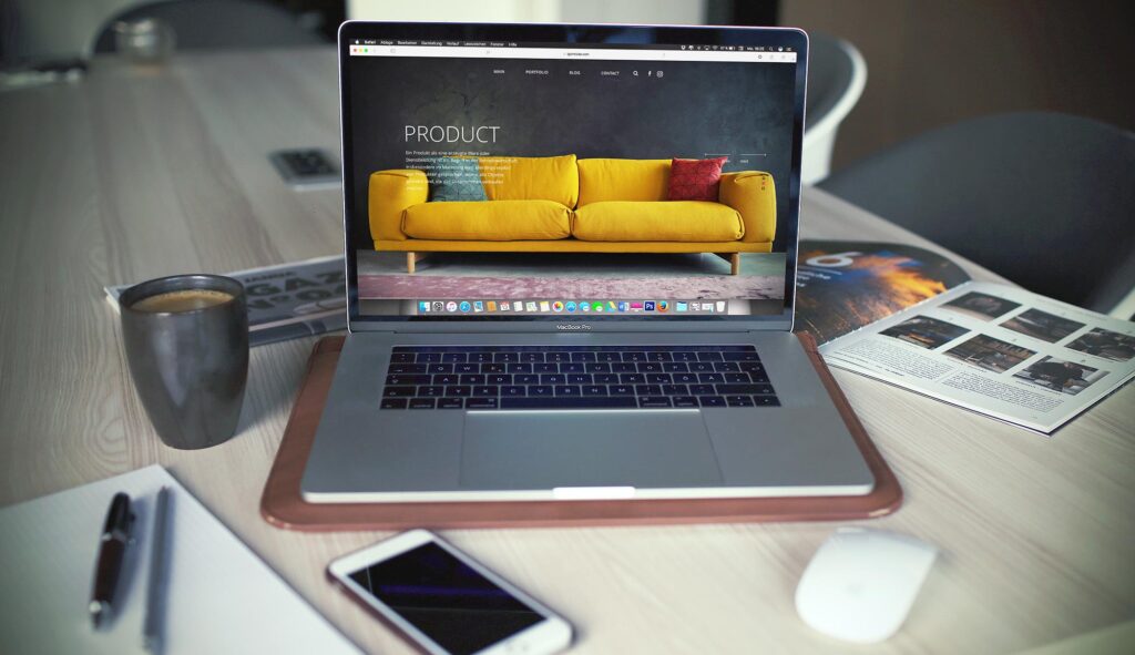 A laptop with a furniture website on the screen sitting on a table with a cup of coffee, iPhone, notebook, book and mouse near by