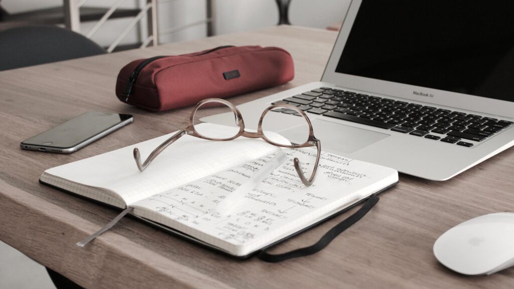 A notebook with a pair of glasses and a laptop on a desk